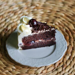 Close-up of a slice of Black Forest cake with cream, cherry, and chocolate on a woven background.