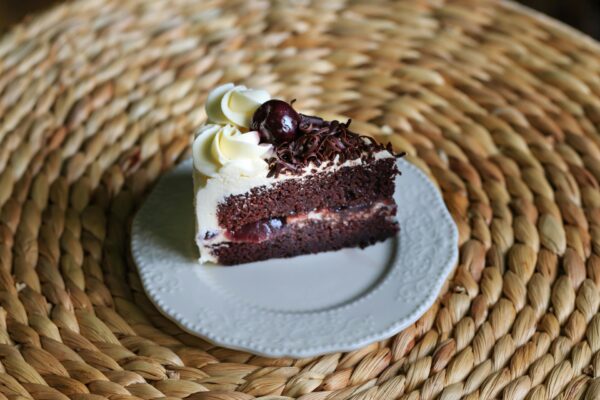 Close-up of a slice of Black Forest cake with cream, cherry, and chocolate on a woven background.
